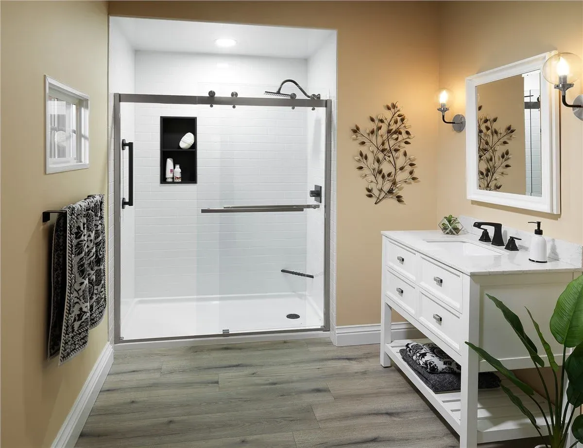 Modern bathroom with glass shower, white tile, beige walls, and wood flooring. A white vanity and a wall mirror with side lighting enhance elegance.