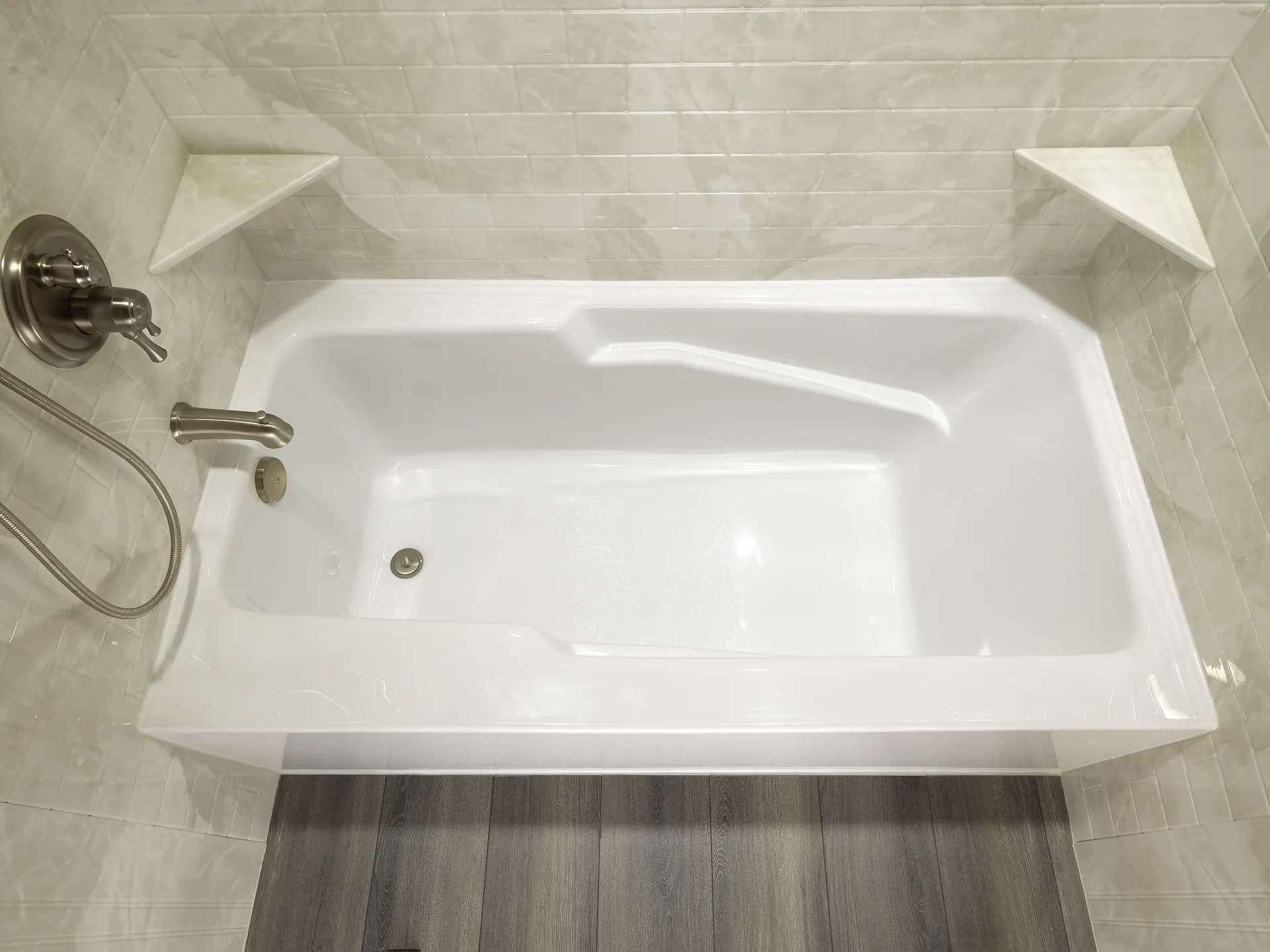 Modern white bathtub with a brushed nickel faucet and handheld showerhead set against glossy beige tiled walls and a dark wood floor.