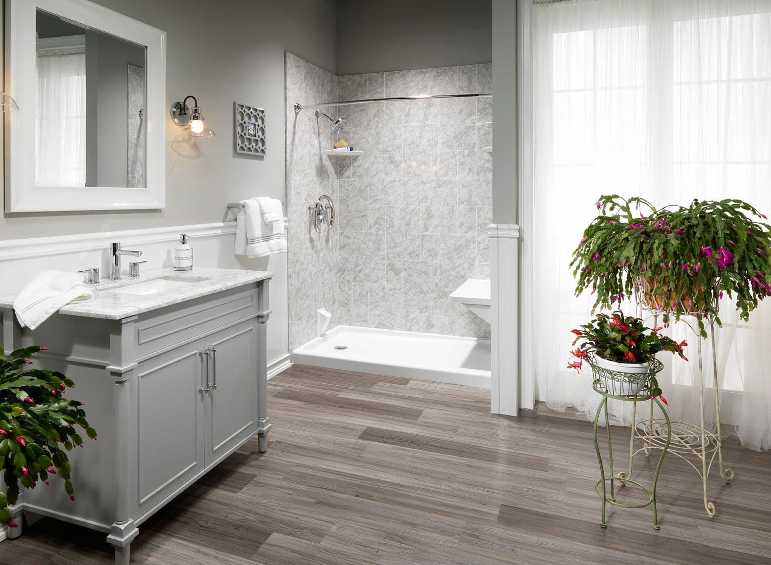 Bright bathroom with gray vanity, marble countertop, and spacious shower. Wooden floor, plants in corners, and soft natural light through sheer curtains.