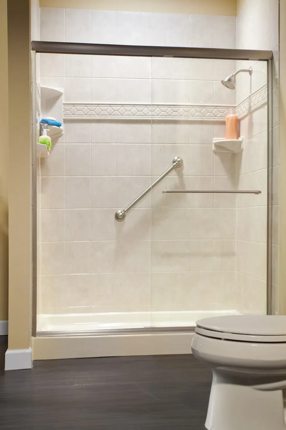Modern bathroom with a tiled walk-in shower featuring safety bars and a showerhead. There's a corner shelf with toiletries and a white toilet nearby.