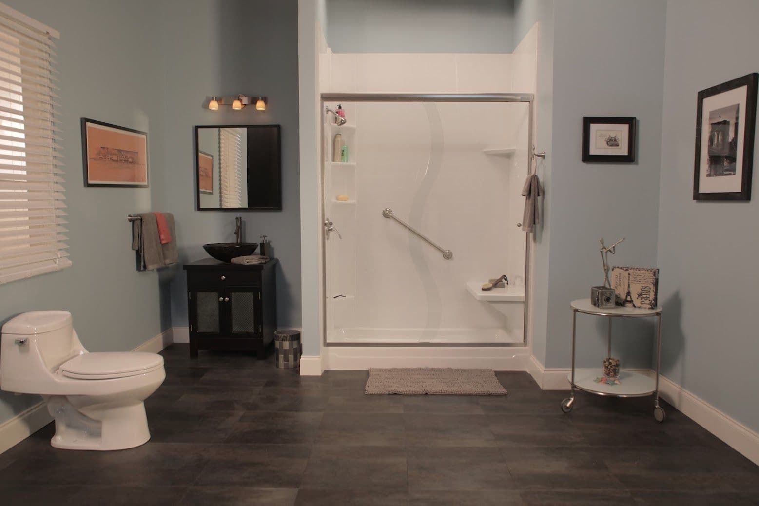 A modern bathroom with a white toilet, large walk-in shower, and black vanity. Light blue walls, framed art, and warm lighting create a calm atmosphere.
