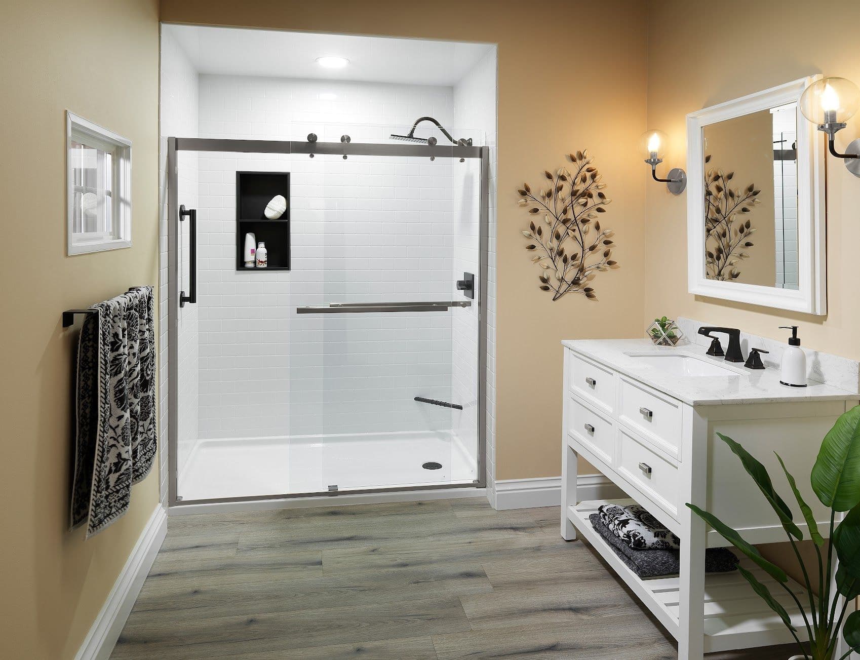 Modern bathroom with beige walls, featuring a glass shower, white vanity, black fixtures, decorative metal wall art, and wooden floor.