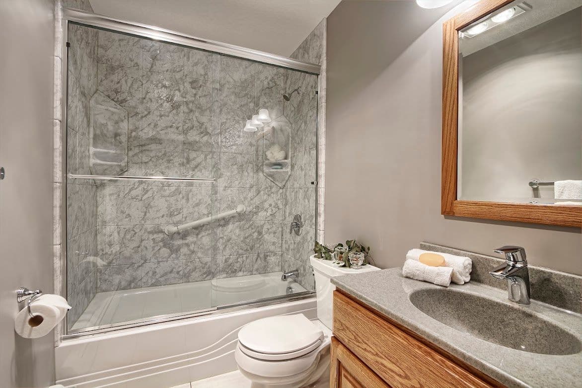 Modern bathroom with gray marble shower, glass sliding doors, white toilet, wooden vanity with gray countertop, and a wooden-framed mirror.