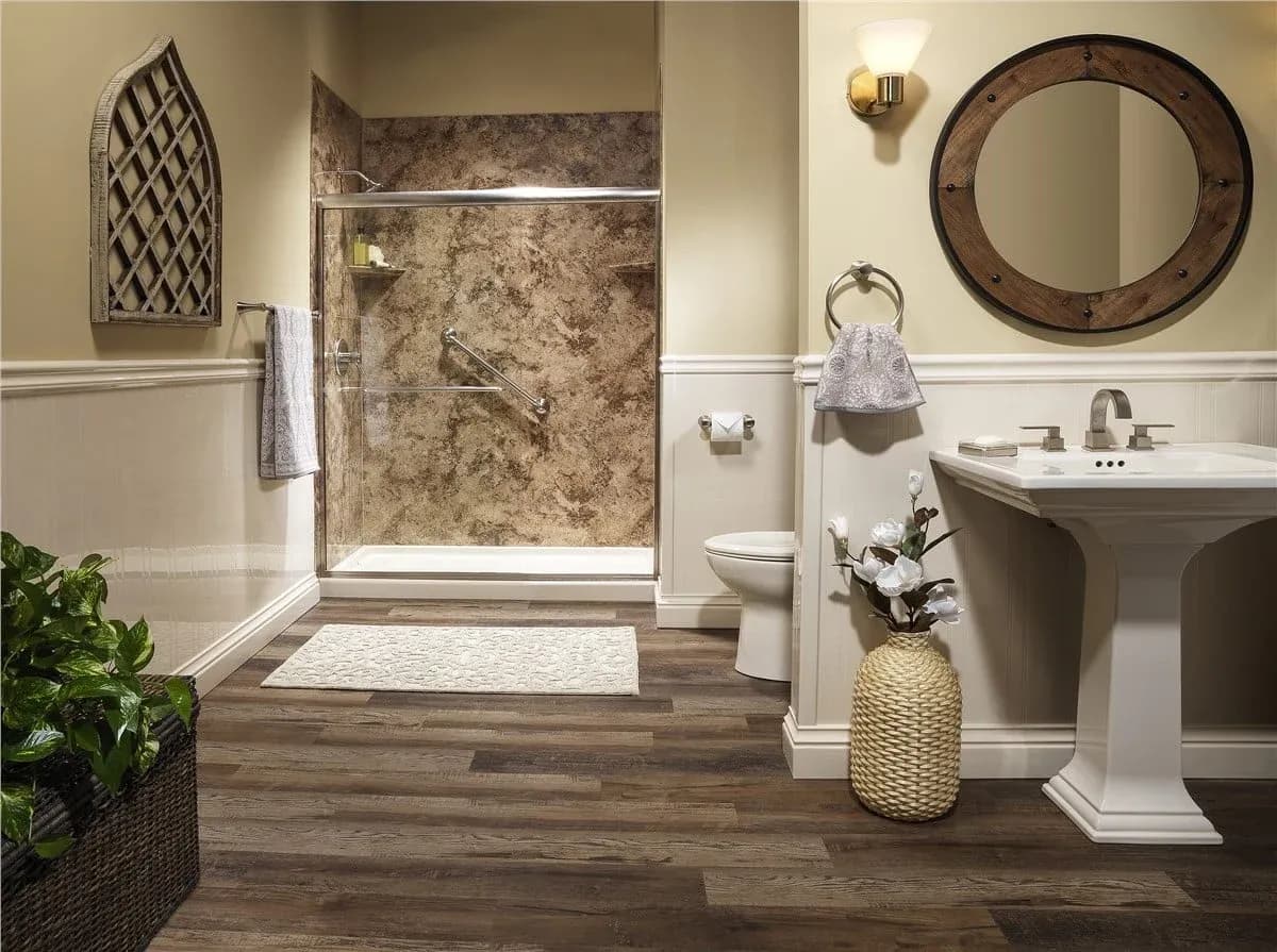 Modern bathroom featuring a glass-enclosed shower with marble walls, a white pedestal sink, wooden floor, decorative plants, and a round mirror.