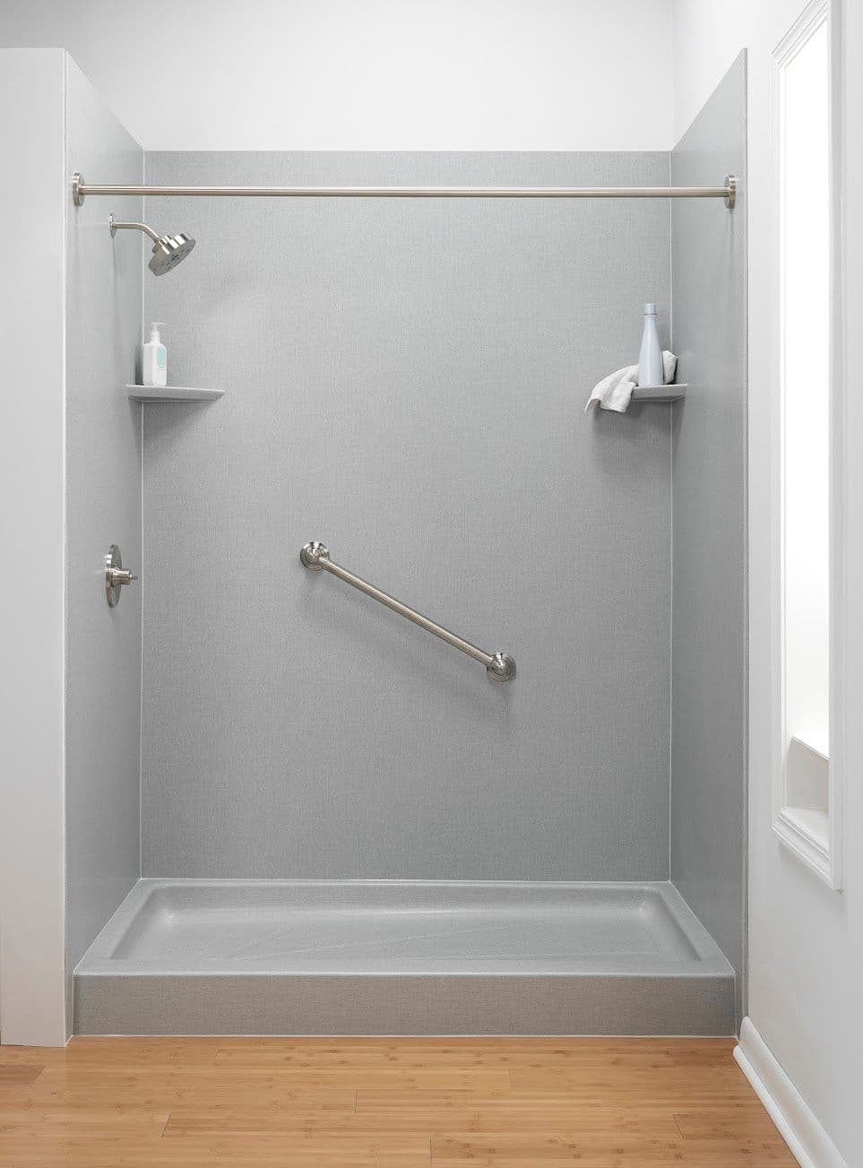Minimalist grey walk-in shower with a metal grab bar, showerhead, and two corner shelves holding toiletries. Light wood floor adds warmth.