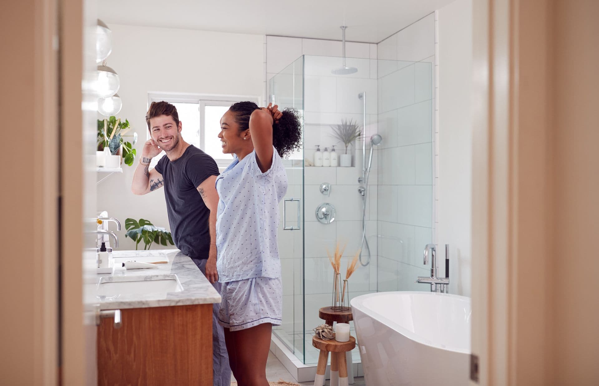 A couple in pajamas stands in a modern bathroom with a glass shower. The woman ties her hair, and both are smiling, conveying a light, cheerful mood.