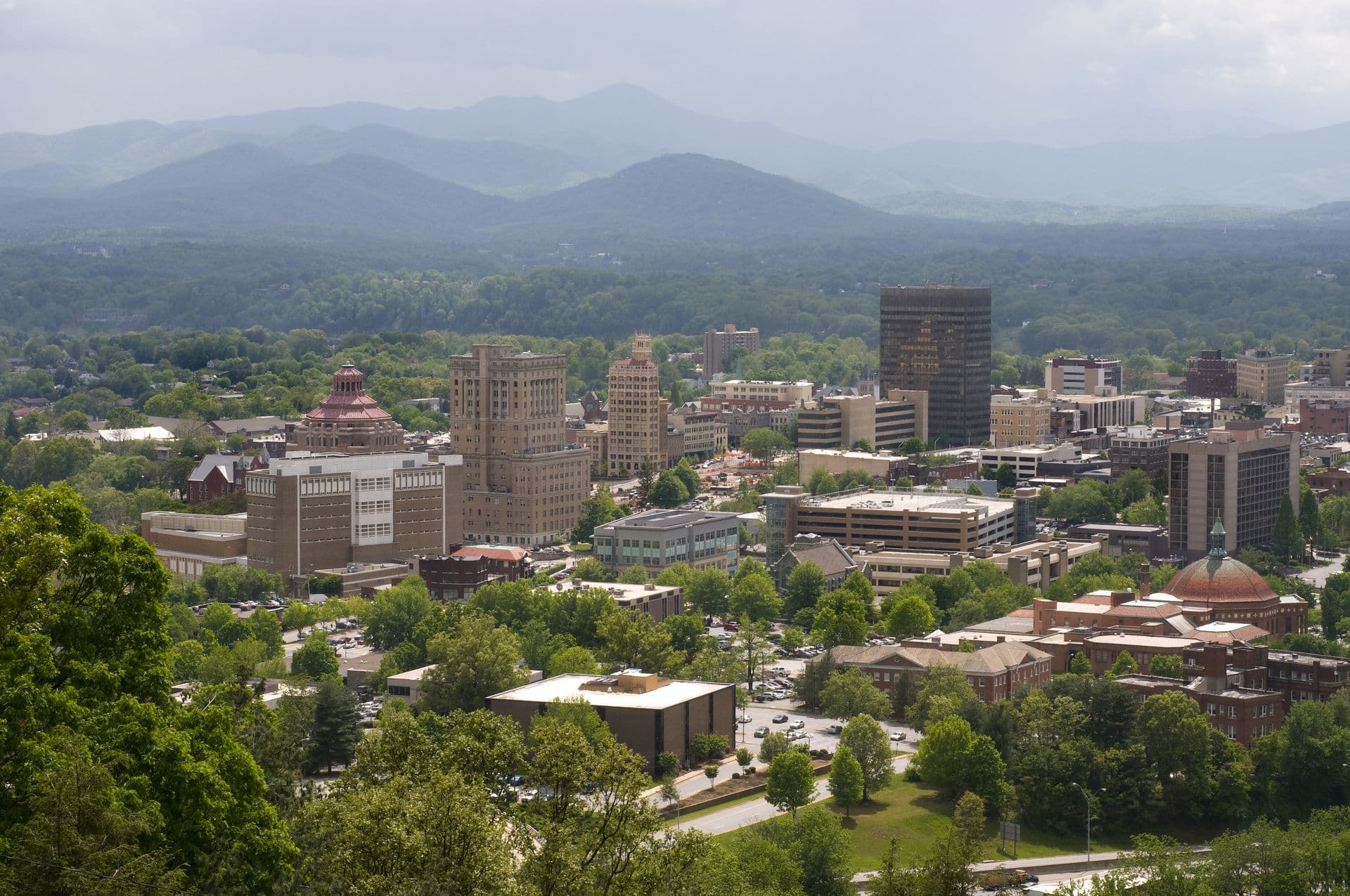 Cityscape of Asheville, North Carolina, surrounded by lush green trees, with tall buildings and distant blue mountains under an overcast sky.