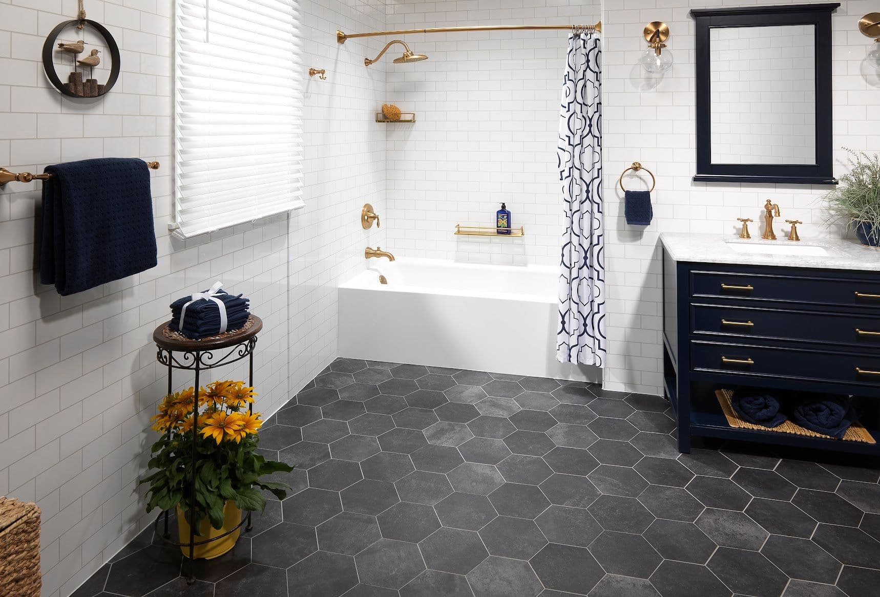 A modern bathroom with white subway tiles and a hexagonal dark floor. It features a white bathtub with gold fixtures, a navy vanity with a marble countertop, and a mirror. Navy towels and a geometric shower curtain add contrast. A plant with yellow flowers adds a touch of nature.