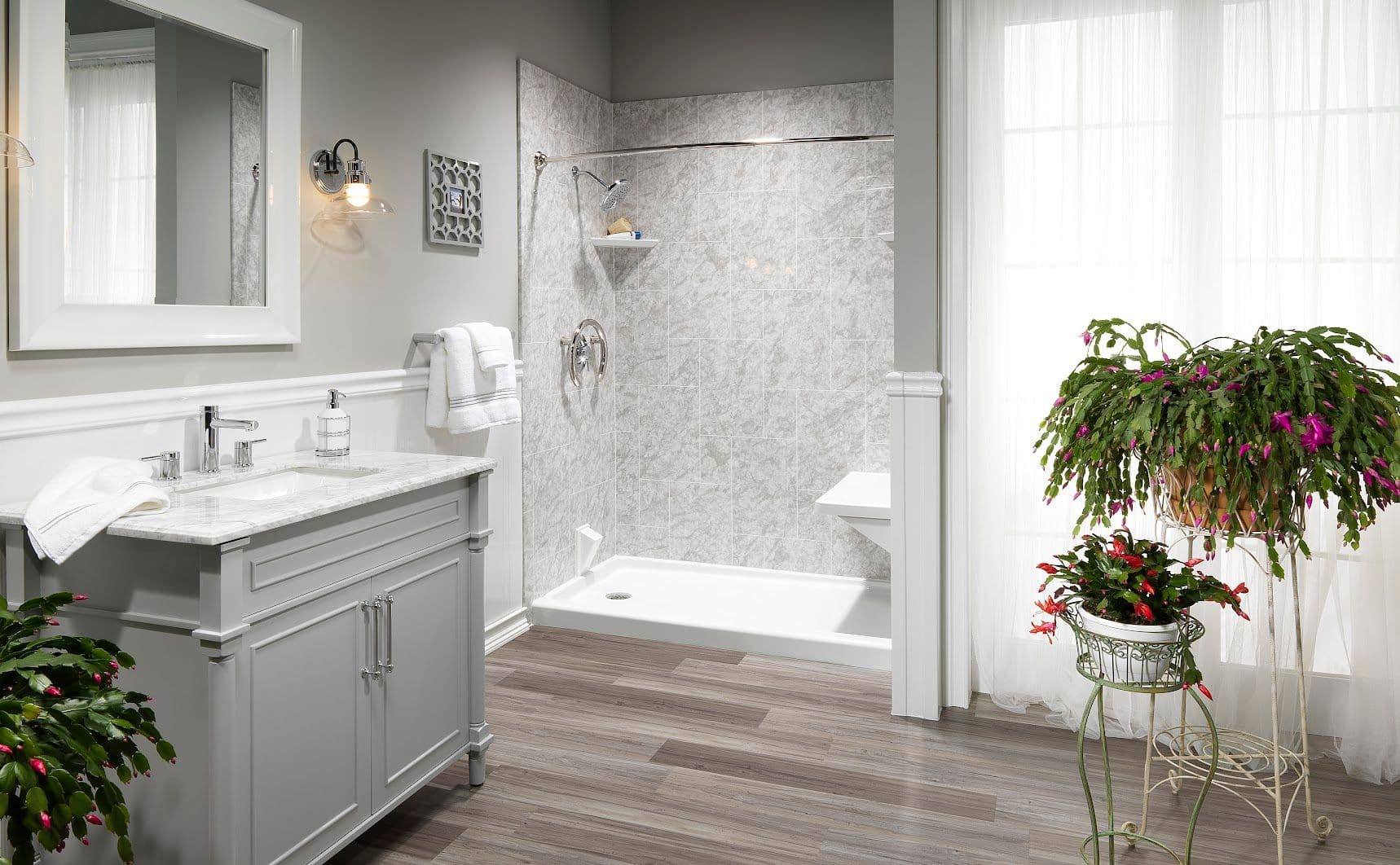 Bright bathroom with a marble-tiled shower, gray vanity with white countertop, wooden floor, and lush potted plants, creating a fresh, serene vibe.
