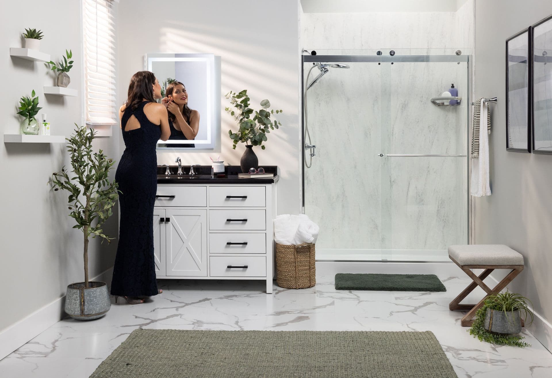 A woman in a black dress admires herself in a bathroom mirror. The space features marble floors, potted plants, a glass shower, and a cozy, modern vibe.
